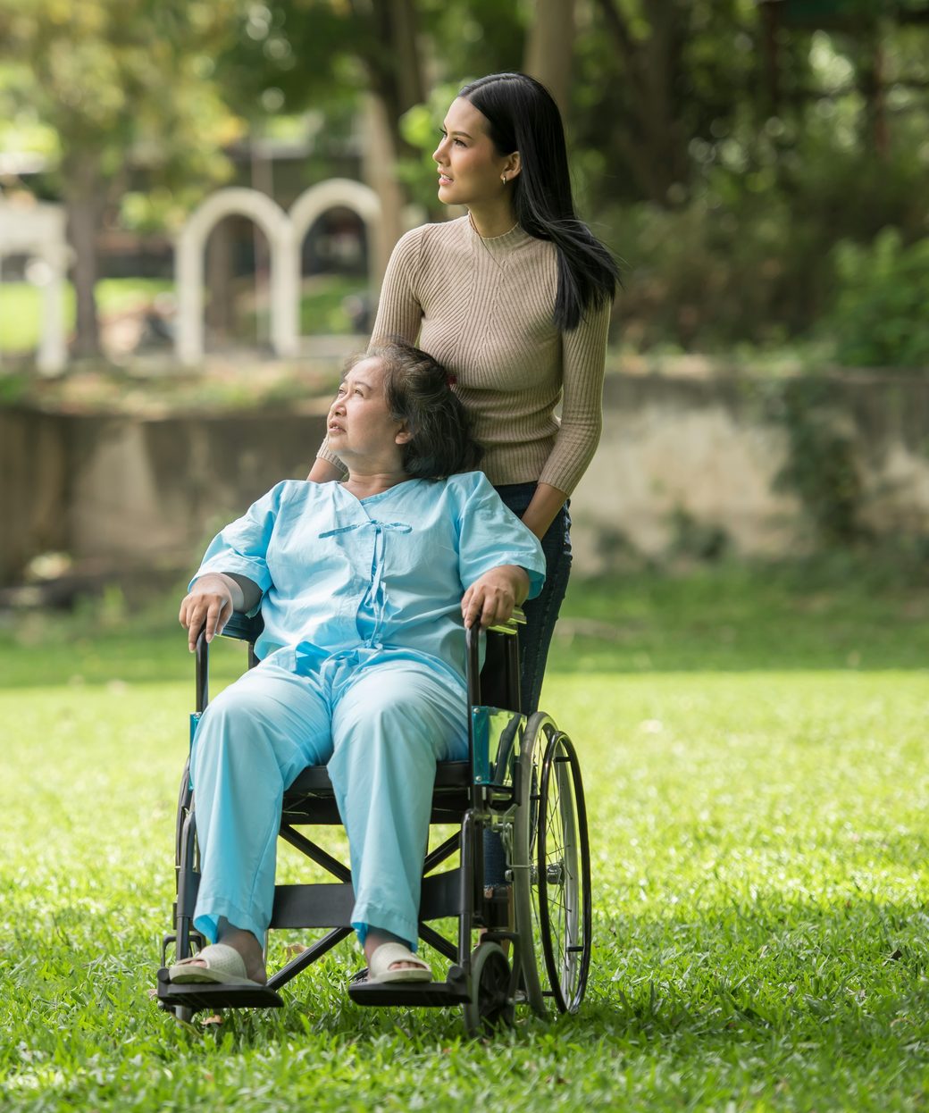 A support worker walking with an older woman in a wheelchair through a sunny park.